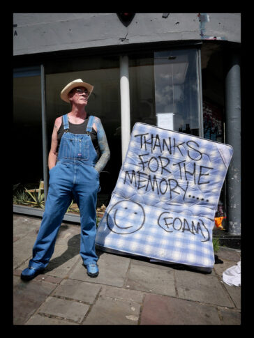 A man in a cowboy hat and denim dungarees - Mr Tony Fortune - stands proudly next to an abandoned mattress on a city sidewalk. The blue and white checked mattress has "Thanks for the memory (foam)" spraypainted across it.